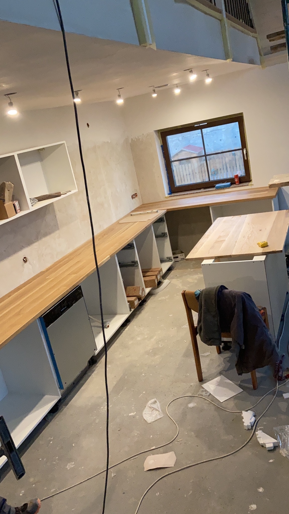 Montaż blatu drewnianego na szafkach kuchennych w surowym stanie, widoczne elementy instalacji elektrycznej i narzędzia na podłodze.