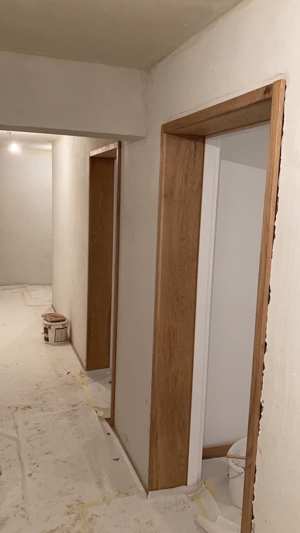 Montaż drewnianych ościeżnic drzwiowych w surowym wnętrzu, widoczny kubeł z farbą i zabezpieczenia podłogi.