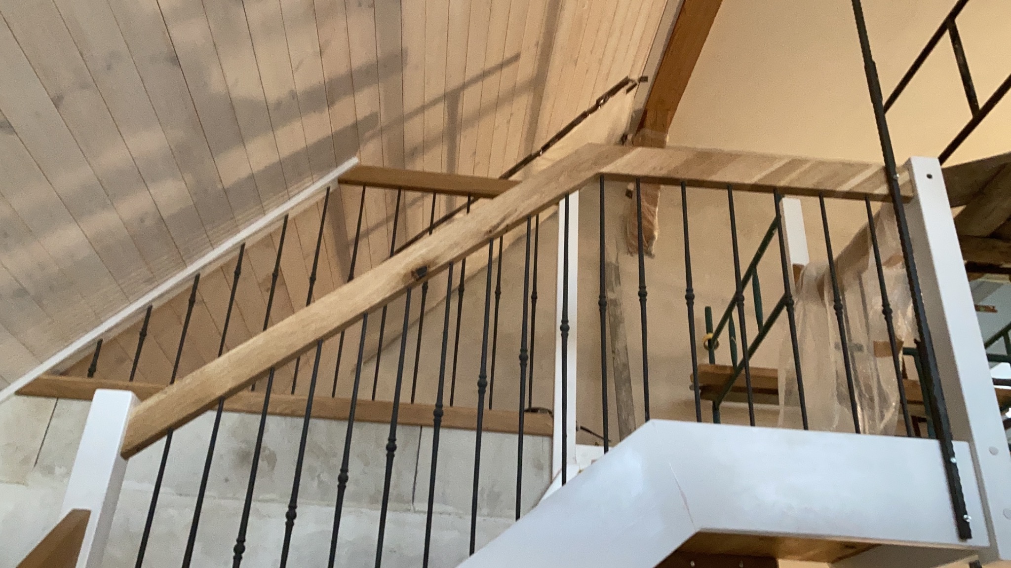 Wnętrze w trakcie budowy: drewniane schody z czarnymi, metalowymi balustradami, białe słupki, surowy drewniany strop, zabezpieczone folią elementy.
