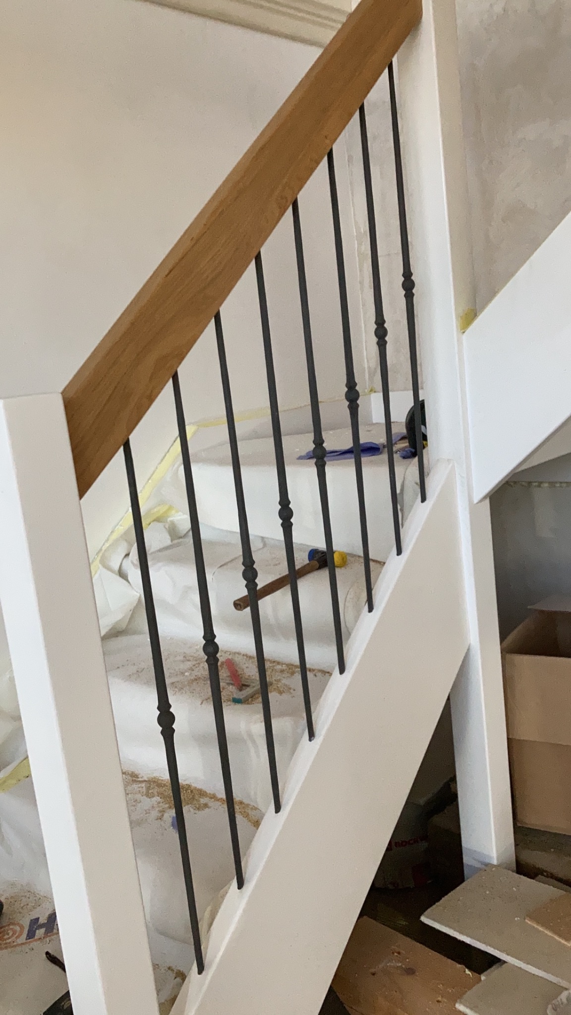 Fragment schodów z białymi stopniami zabezpieczonymi folią, drewnianą poręczą i czarnymi, kutymi balustradami w trakcie montażu, widoczne narzędzia i drobne śmieci.