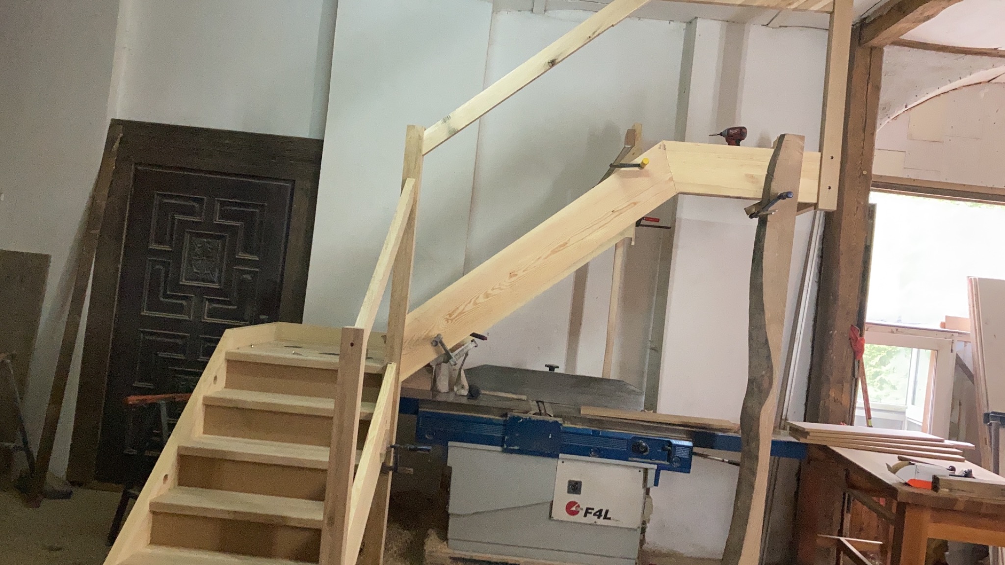 Wnętrze warsztatu z widoczną konstrukcją drewnianych schodów w trakcie montażu, z użyciem wiertarki akumulatorowej i zacisków stolarskich, z maszyną stolarską w tle i starą, ozdobną, ciemną futryną...