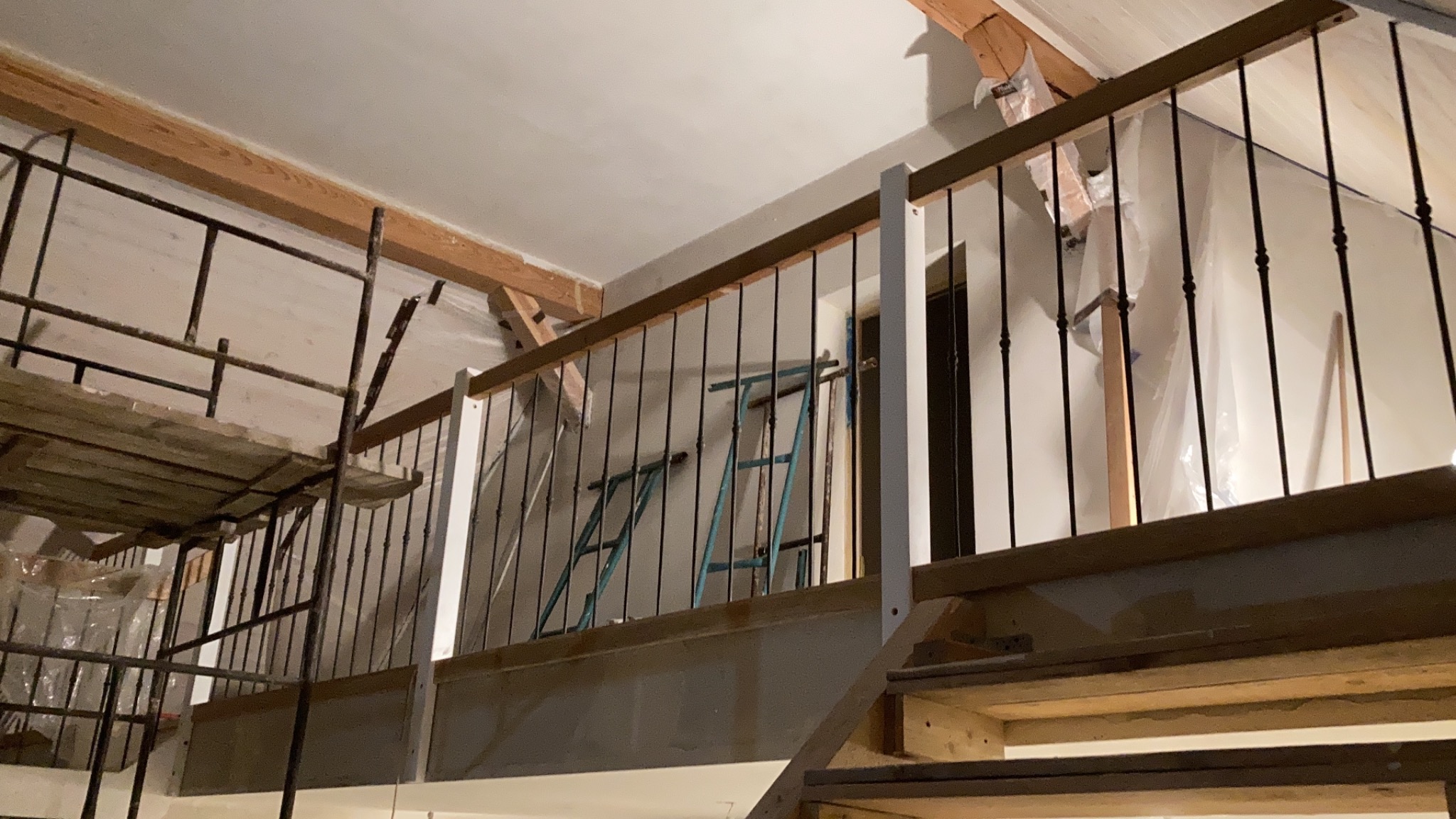 Wnętrze z drewnianymi schodami w trakcie montażu, widoczne rusztowanie i zabezpieczenia folią malarską, barierka z czarnymi prętami i drewnianym poręczą.