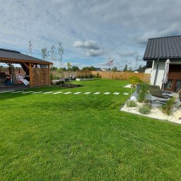 AMADEUSZ PIETRZAK FIDELIUS - Zadbany trawnik z ułożonymi płytami chodnikowymi, drewniana altana z huśtawką, ogrodzenie z desek i rabaty z białym kamieniem, widok na nowoczesny dom z szarą dachówką.