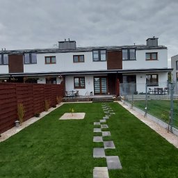 AMADEUSZ PIETRZAK FIDELIUS - Widok ogrodu przed szeregową zabudową, z równo przystrzyżonym trawnikiem, ścieżką z kwadratowych płyt, młodym drzewkiem w centralnej części, brązowym drewnianym ogrodzeniem po lewej i metalowym po...