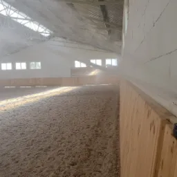 Automatyczny system nawadniania w hali jeździeckiej, rozpylający wodę na piaszczystą nawierzchnię, widoczny z bliska czarny zraszacz zamontowany na drewnianej bandzie przy ścianie z betonu...