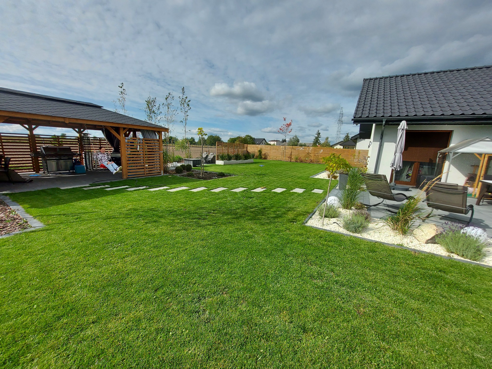 Zadbany trawnik z ułożonymi płytami chodnikowymi, drewniana altana z huśtawką, ogrodzenie z desek i rabaty z białym kamieniem, widok na nowoczesny dom z szarą dachówką.