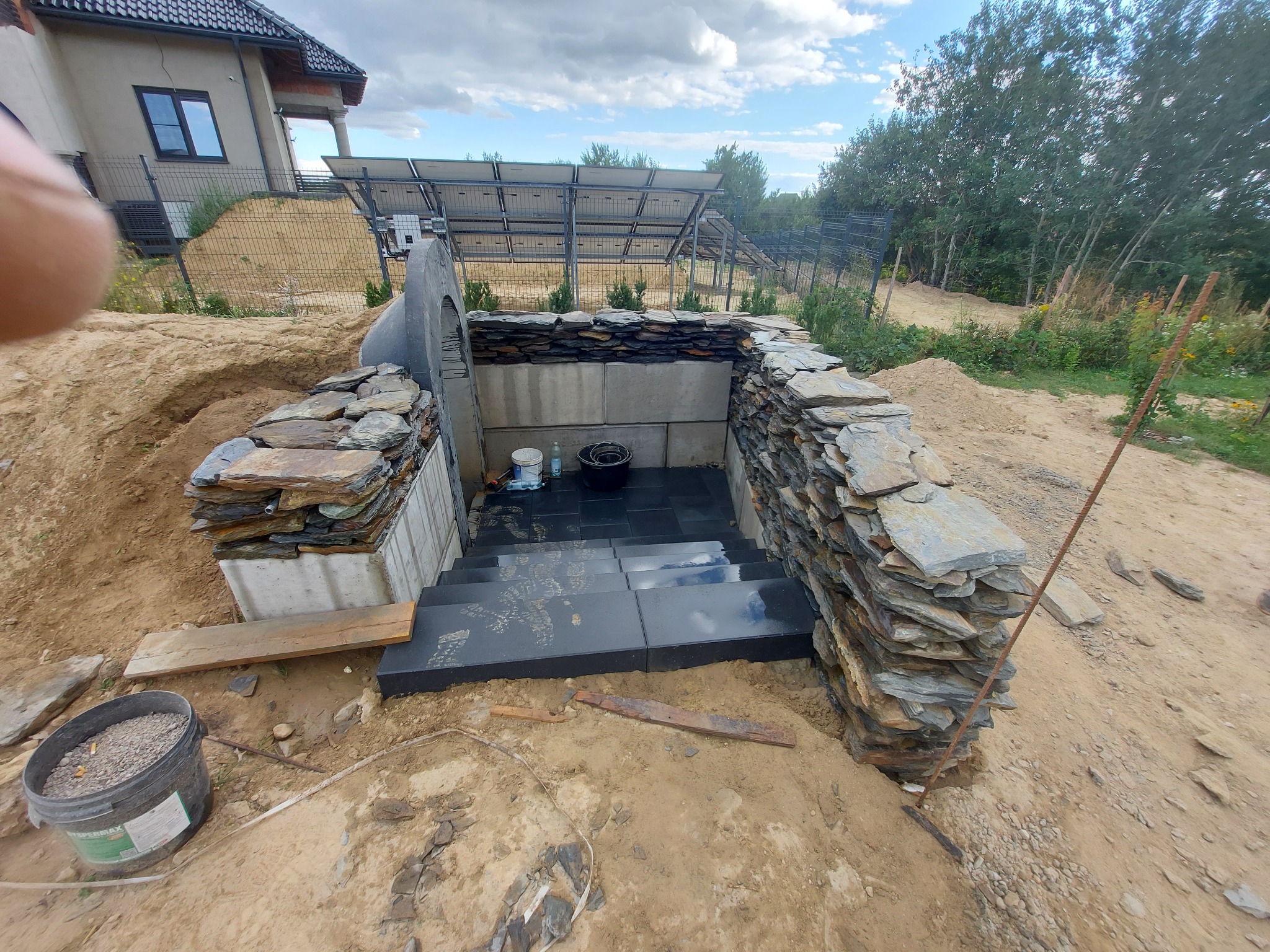 Schody zewnętrzne wyłożone czarnym kamieniem prowadzące do piwnicy, otoczone murem z łupka, w tle dom i panele słoneczne, teren w trakcie prac ziemnych.