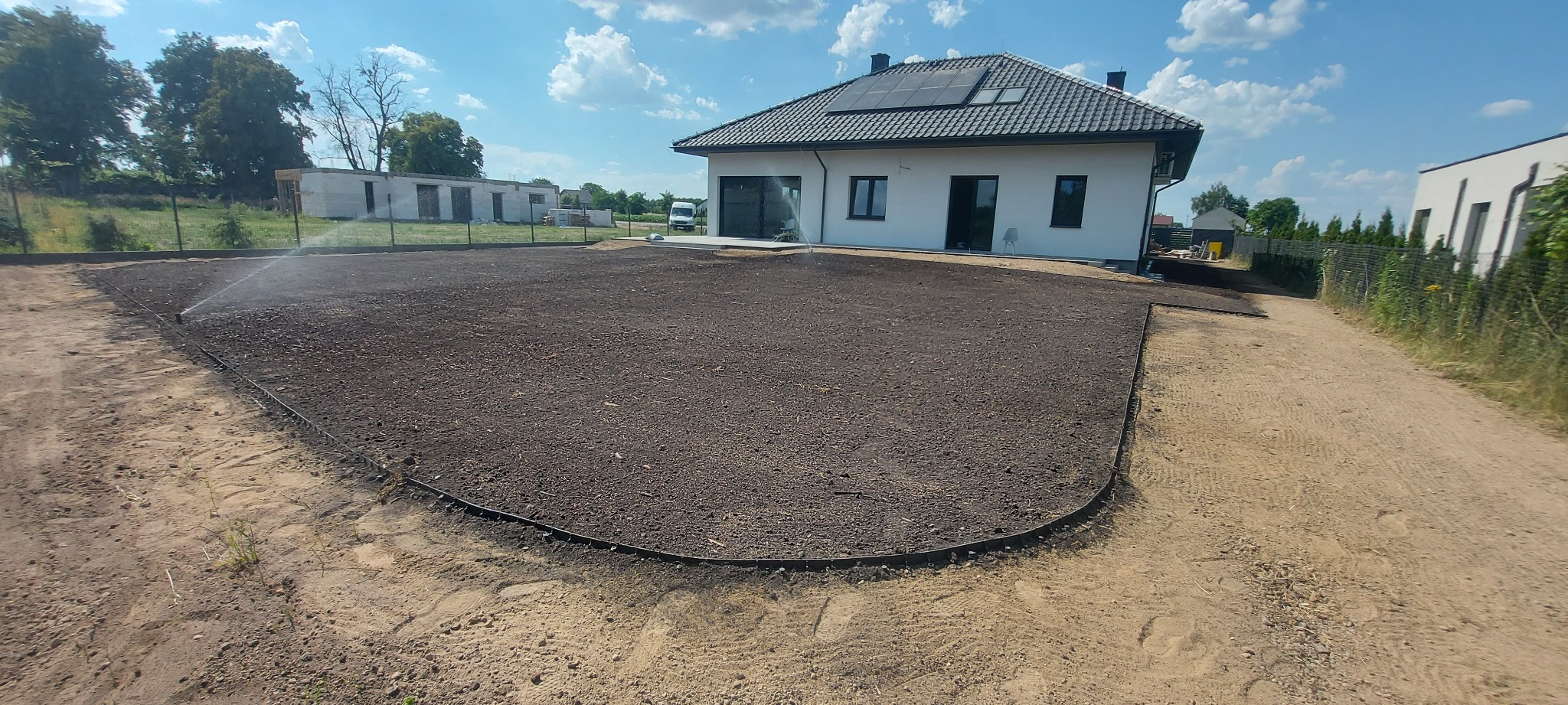 Przygotowany teren pod trawnik z nawadnianiem, obrzeżami i widocznym nowym domem z panelami słonecznymi w tle.