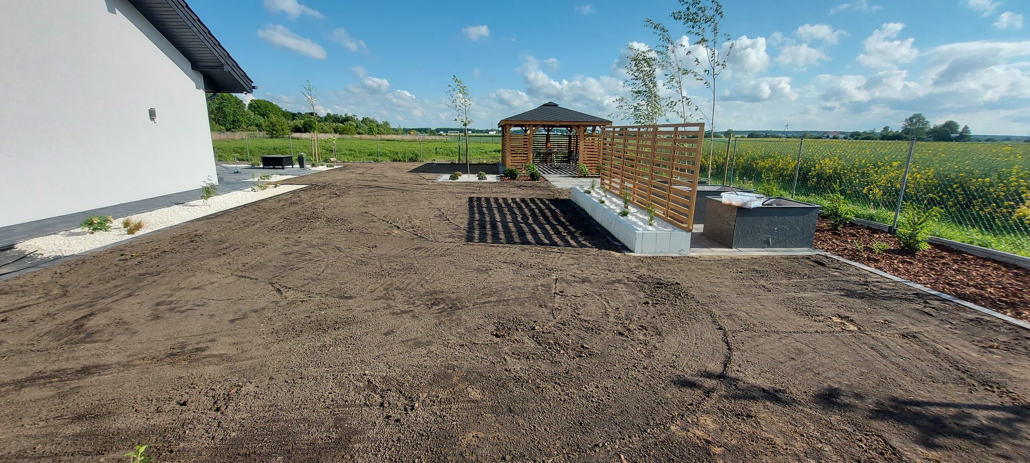 Przygotowany teren pod trawnik z widoczną altaną, rabatami z białym kamieniem i młodymi drzewkami, na tle pola rzepaku.