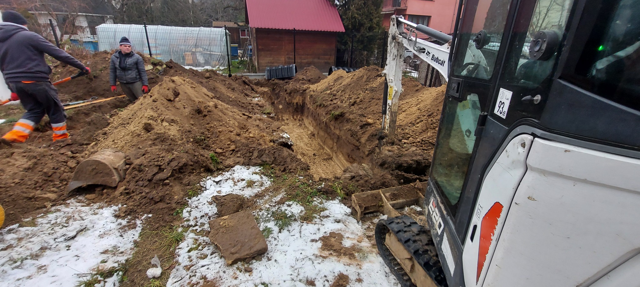Wykop wąskiego rowu w ziemi z resztkami śniegu, wykonany koparką Bobcat E20, z widocznymi pracownikami i stertami ziemi w tle.