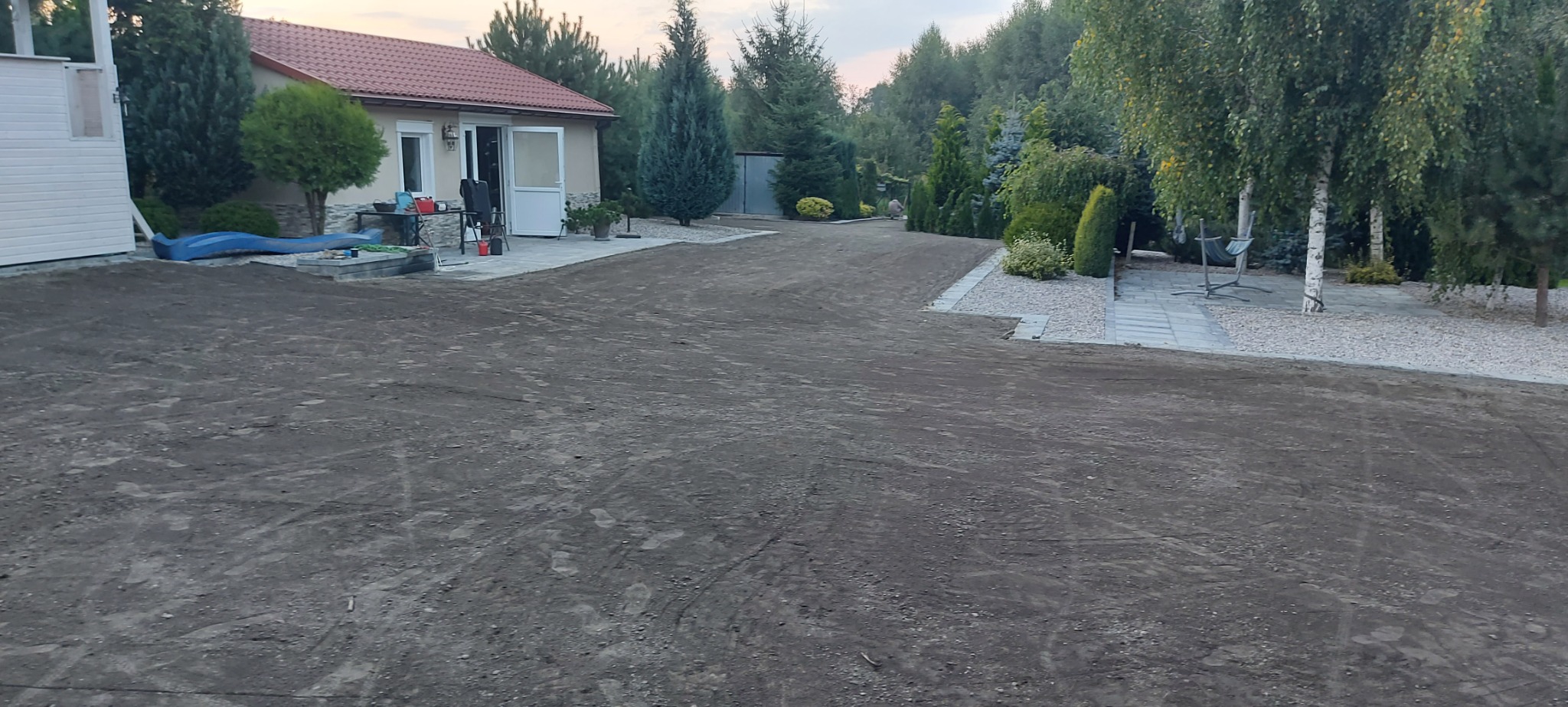 Przygotowany teren pod trawnik z widocznymi śladami po maszynach, obok domu z czerwonym dachem i elementami małej architektury ogrodowej.