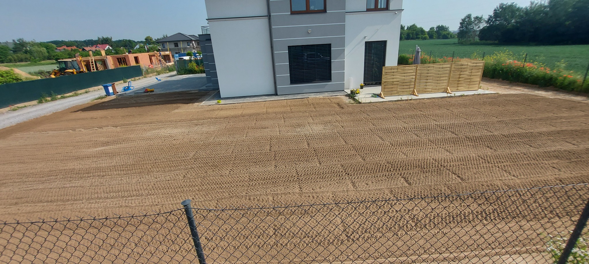 Przygotowany teren pod trawnik, z widocznymi śladami po maszynach, w tle nowy dom z drewnianym ogrodzeniem i budowa, widok zza metalowego ogrodzenia.