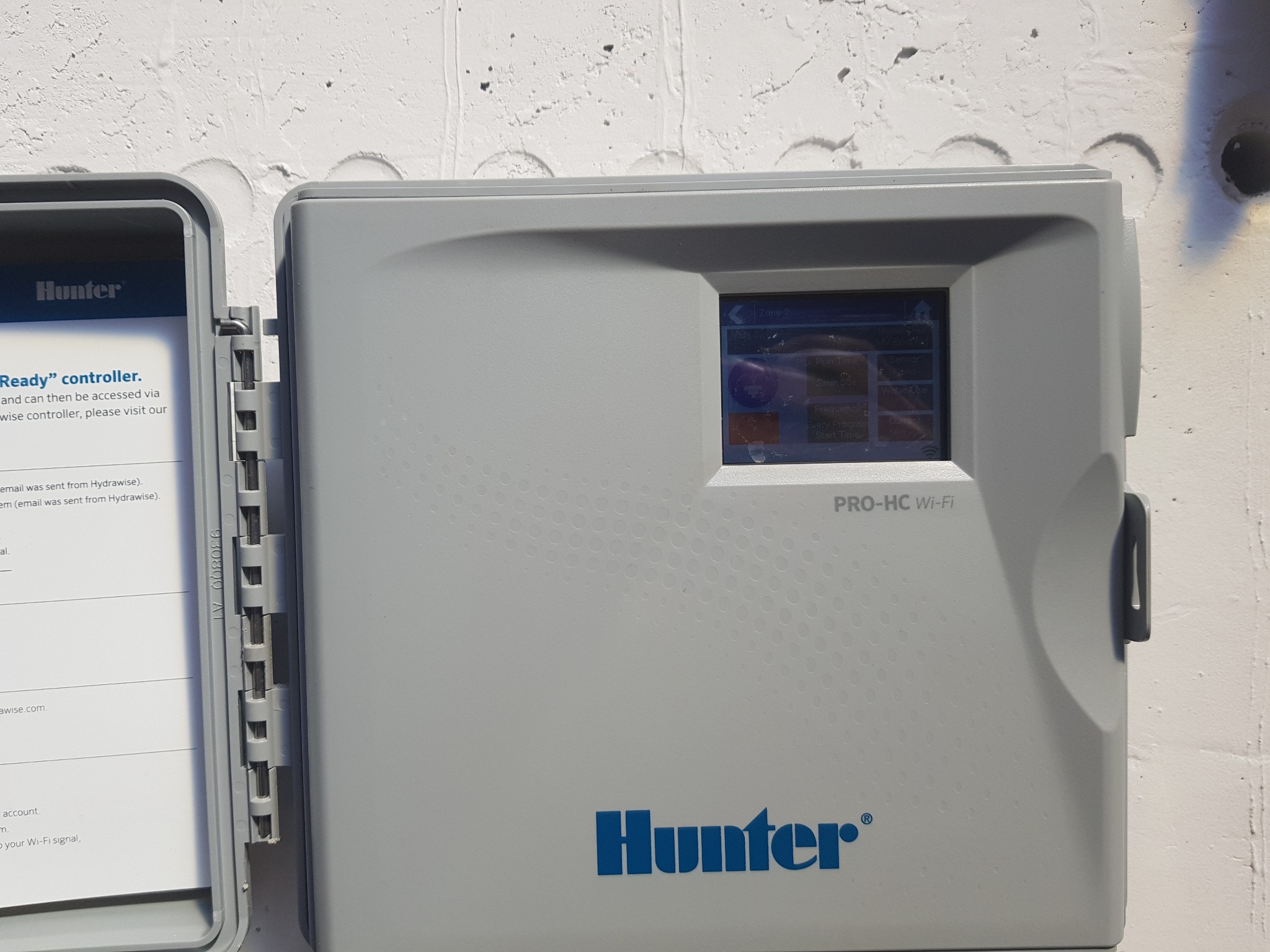 Szary kontroler nawadniania Hunter PRO-HC Wi-Fi zamontowany na białej, teksturowanej ścianie, z widocznym ekranem i otwartą obudową ukazującą instrukcję.