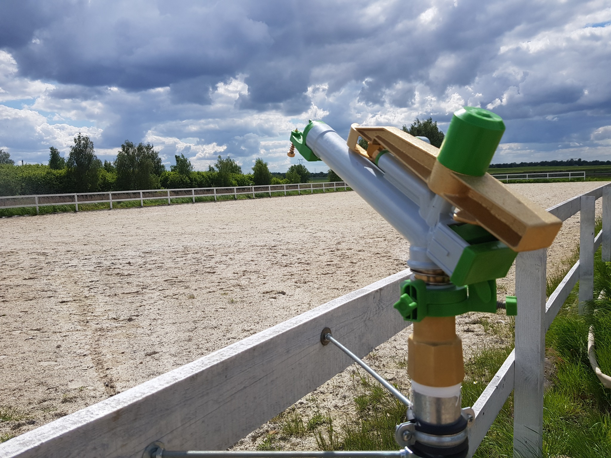 Zraszacze rotacyjne, z elementami w kolorze zielonym i złotym, zamontowane na białym ogrodzeniu, nawadniające piaszczysty teren pod pochmurnym niebem.