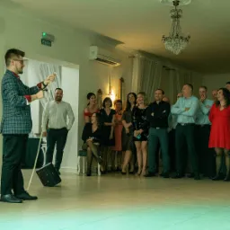 Iluzjonista w kraciastej marynarce wykonuje trik z liną i walizką na tle publiczności w eleganckim wnętrzu sali balowej z żyrandolem.