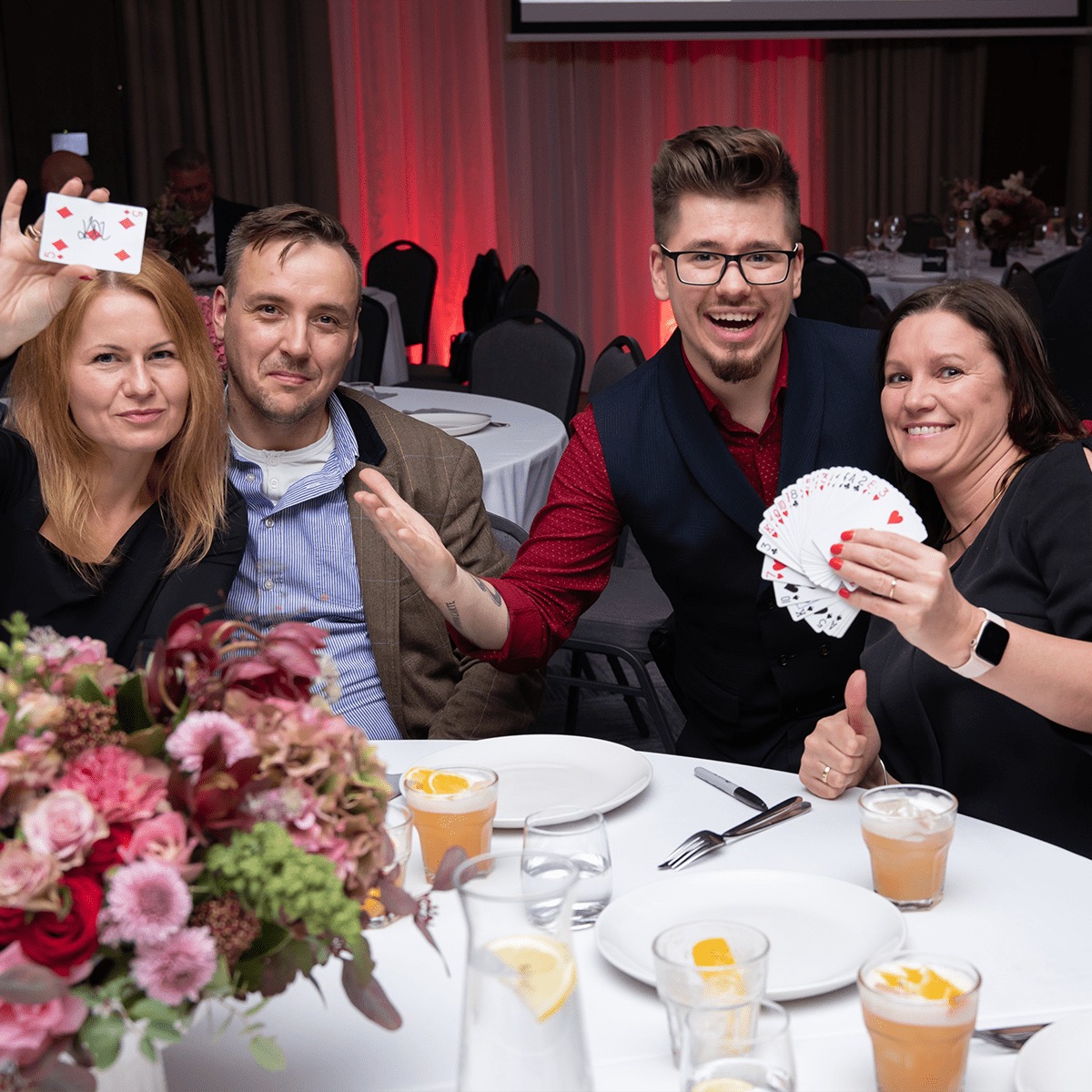 Iluzjonista w czerwonej koszuli i ciemnej kamizelce prezentuje karty publiczności podczas występu na imprezie, otoczony uśmiechniętymi gośćmi przy stole z napojami i kwiatami.