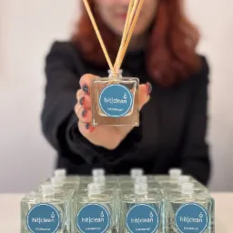 Kobieta prezentuje dyfuzor zapachowy z logo 'hit|clean', szereg identycznych dyfuzorów na białym stole w tle.