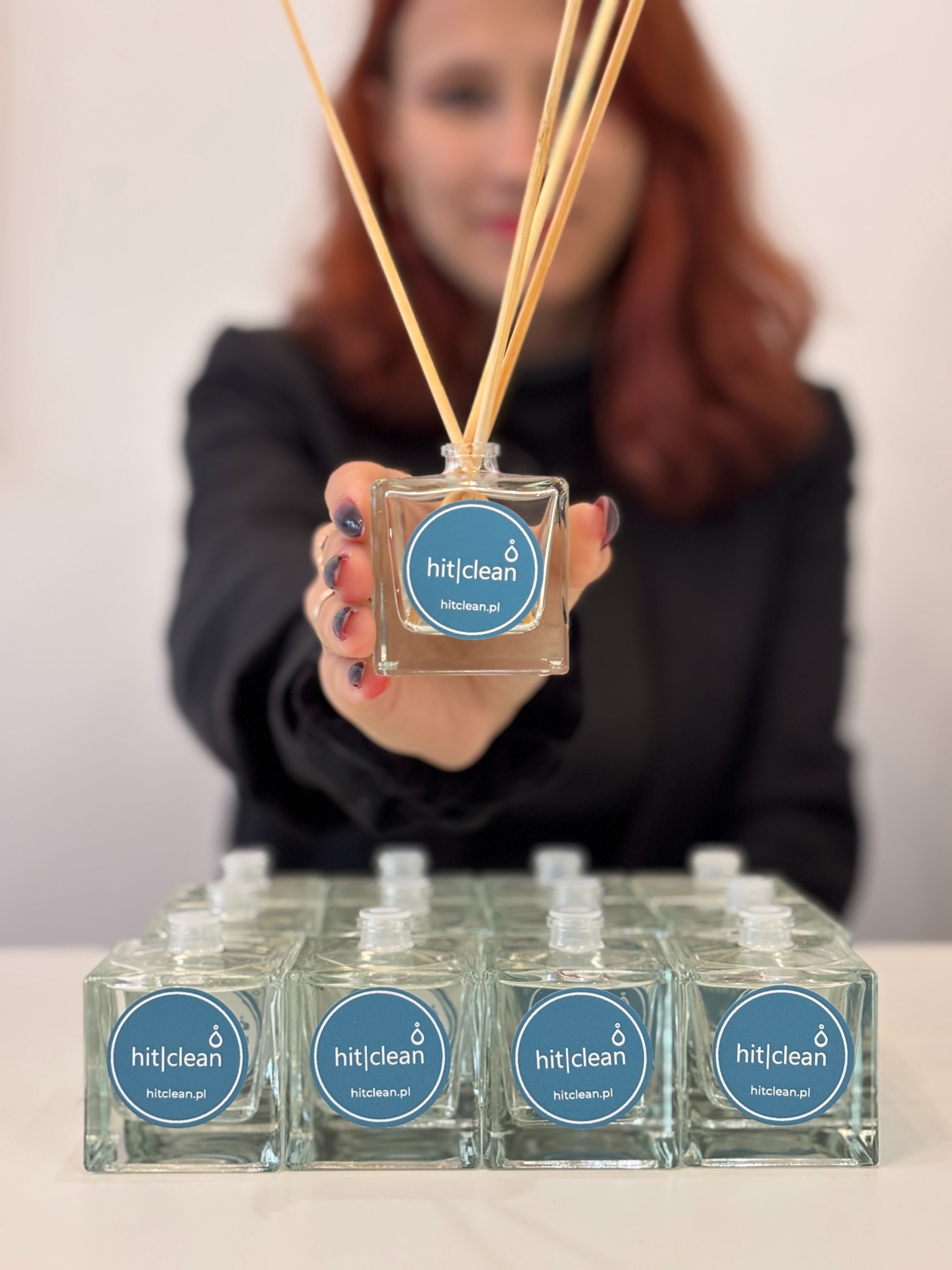 Kobieta prezentuje dyfuzor zapachowy z logo 'hit|clean', szereg identycznych dyfuzorów na białym stole w tle.