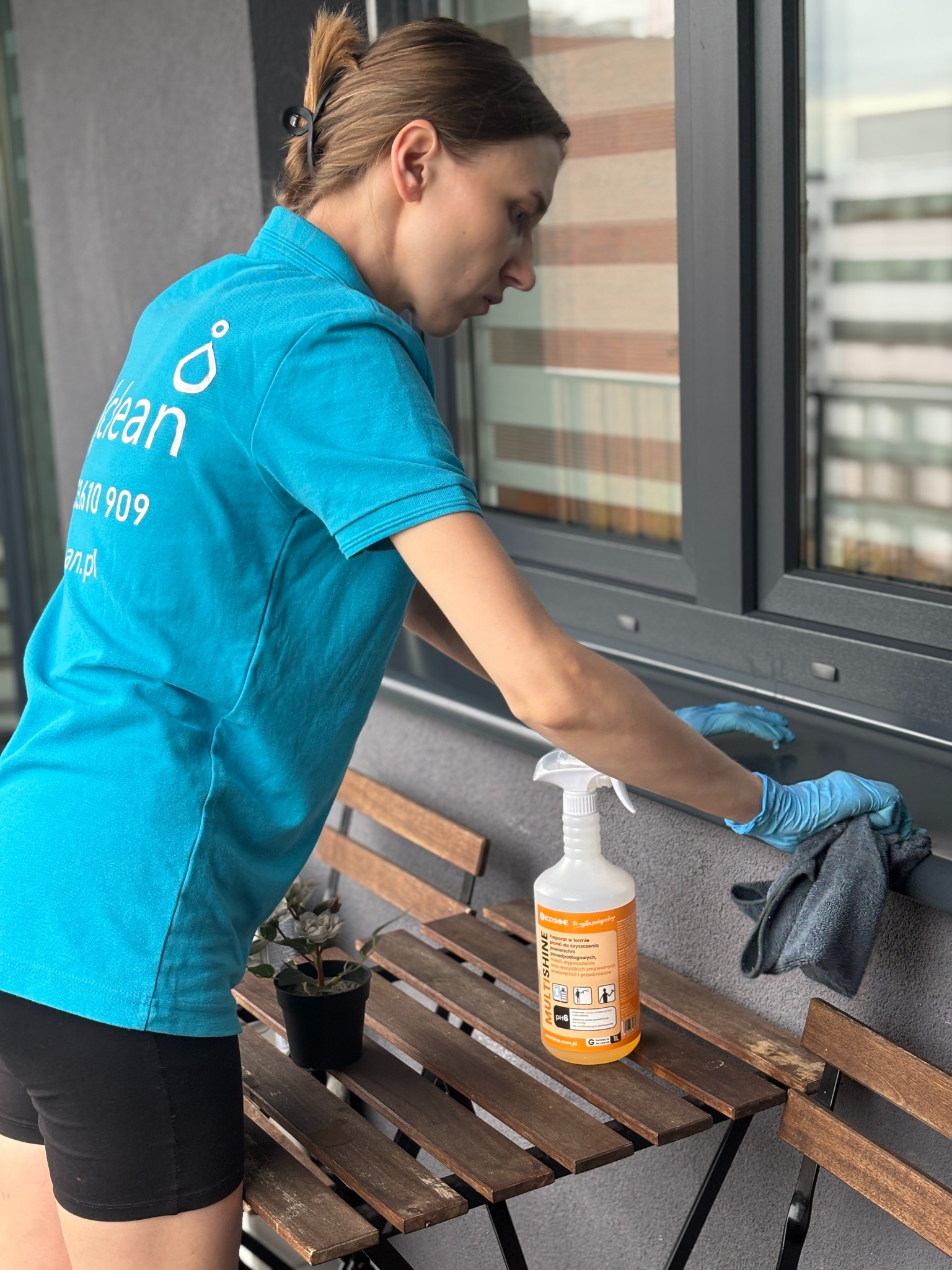 Kobieta w niebieskiej koszulce firmowej czyści szarą ramę okienną na balkonie, używając spryskiwacza i szmatki z mikrofibry, obok stoi drewniany stolik z kwiatkiem w doniczce.