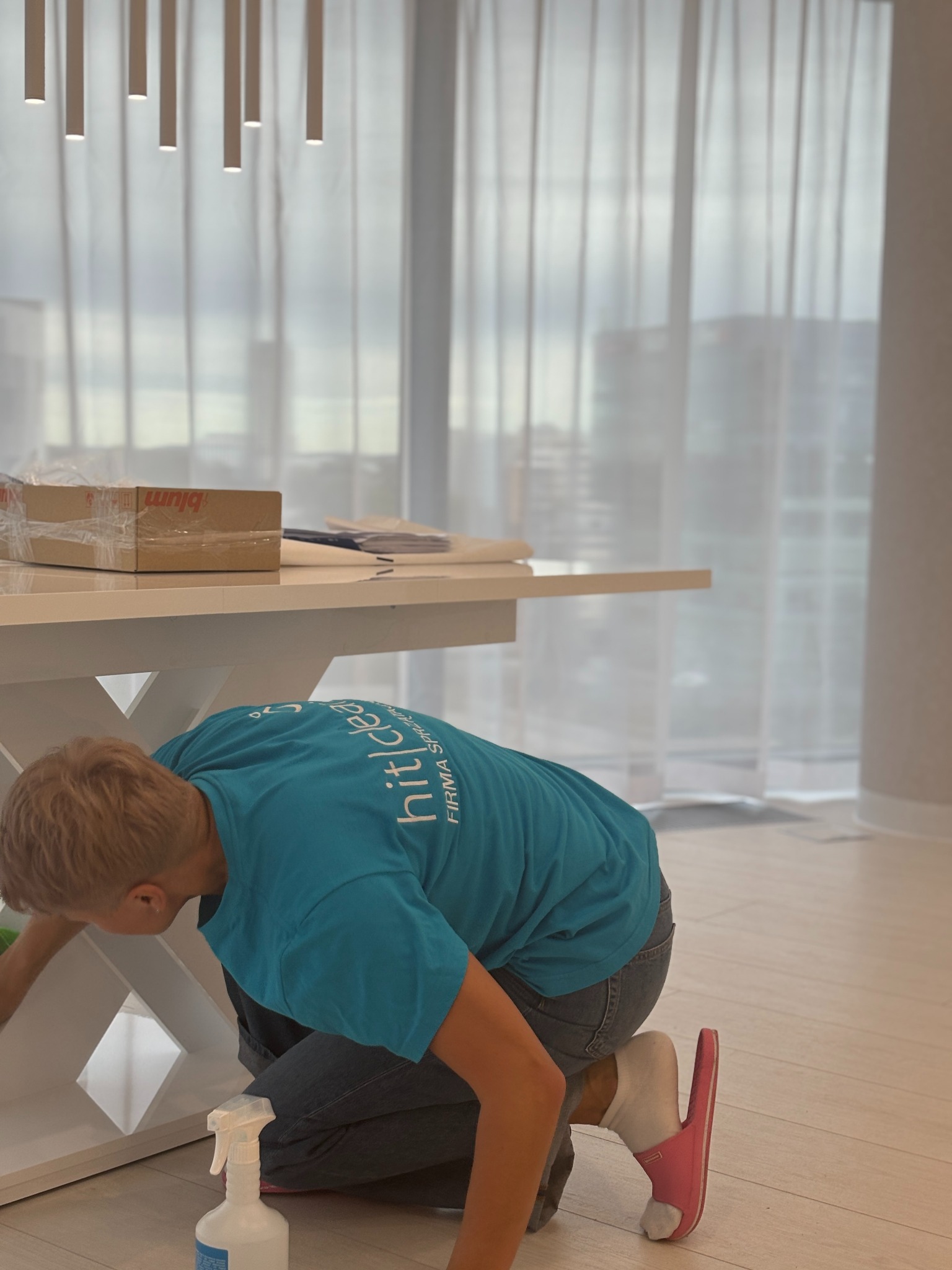 Osoba w niebieskiej koszulce z logo firmy sprzątającej, klęcząca podczas czyszczenia białej designerskiej komody w jasnym wnętrzu z dużym oknem i designerskim oświetleniem sufitowym. Na blacie leżą...