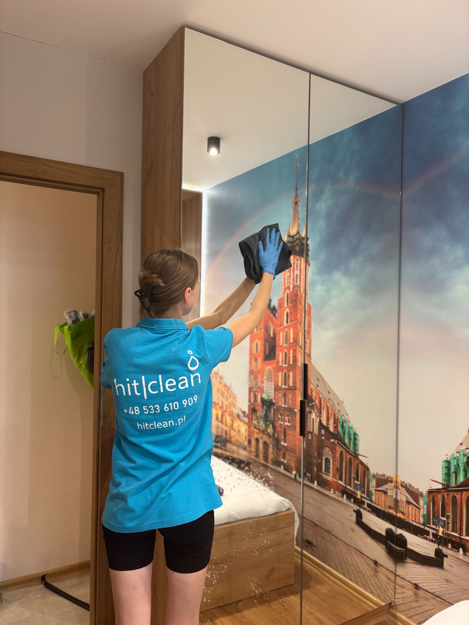 Pracownica w niebieskiej koszulce z logo firmy hit|clean czyści lustrzane drzwi szafy z fototapetą przedstawiającą Bazylikę Mariacką w Krakowie, używając szarej ściereczki i niebieskich rękawiczek.
