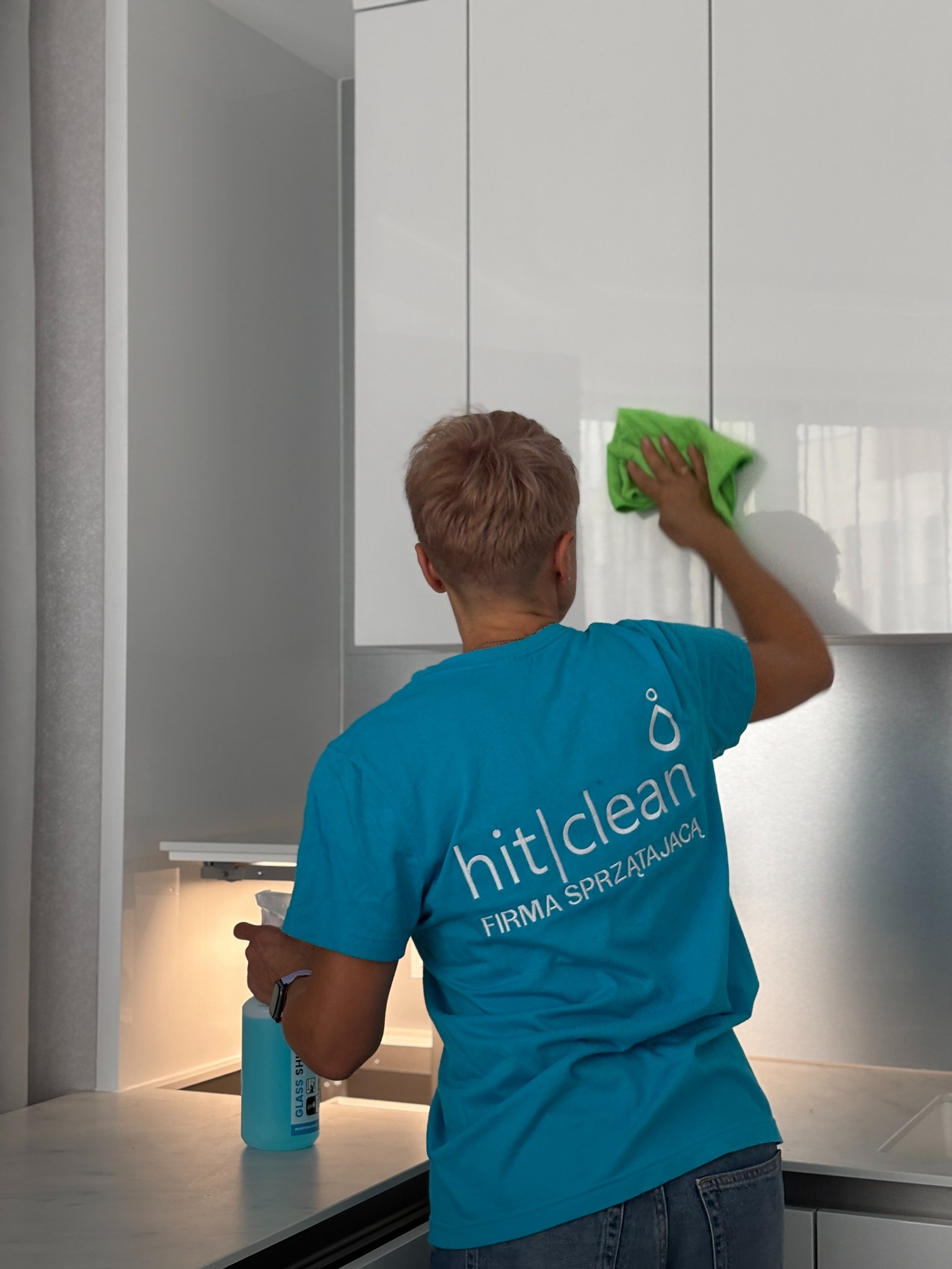 Pracownik firmy sprzątającej w turkusowej koszulce z logo, czyści zieloną ściereczką białe, lakierowane fronty szafek kuchennych, obok stoi butelka środka do czyszczenia szkła.
