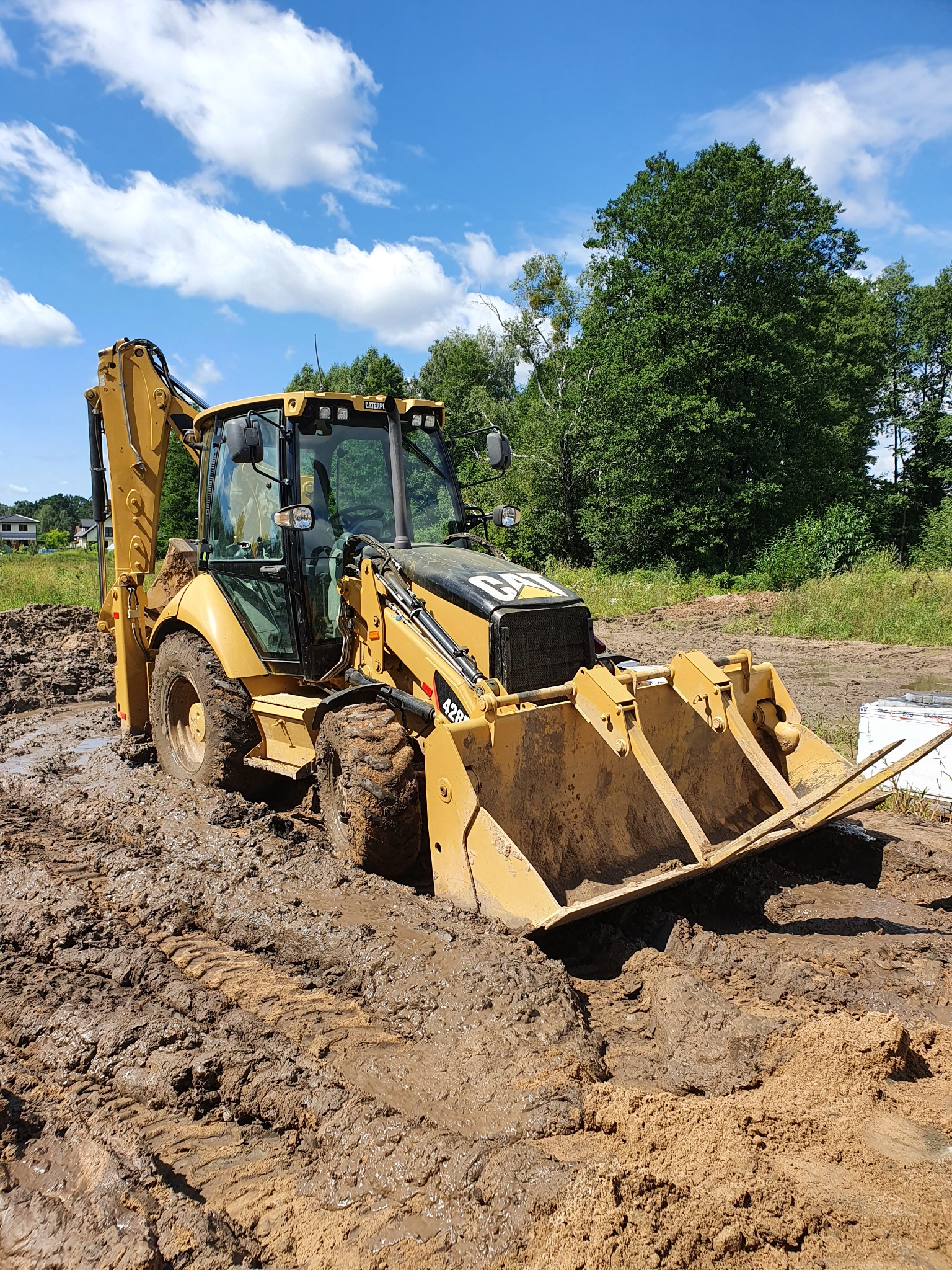 Żółta koparko-ładowarka CAT 428 w błotnistym terenie, widok z przodu na łyżkę z zębami, błękitne niebo z chmurami w tle.
