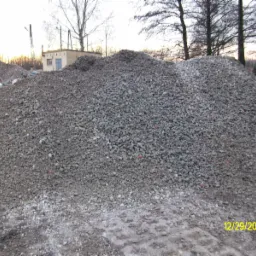 Duża pryzma szarego kruszywa z domieszką cegieł na placu budowy, w tle widoczny budynek i drzewa.