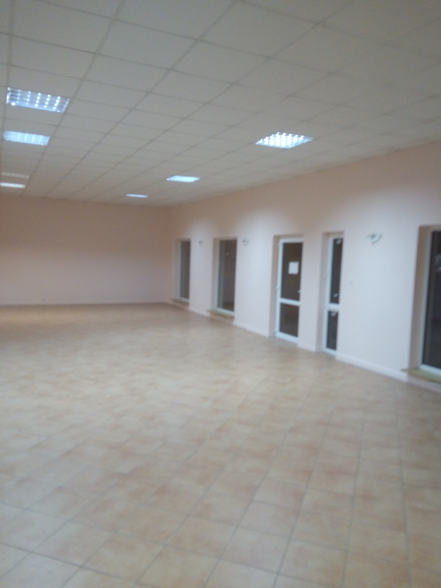 Puste pomieszczenie z beżowymi ścianami, jasnymi płytkami na podłodze i rzędem drzwi z białymi ościeżnicami, oświetlone kasetonowym sufitem.