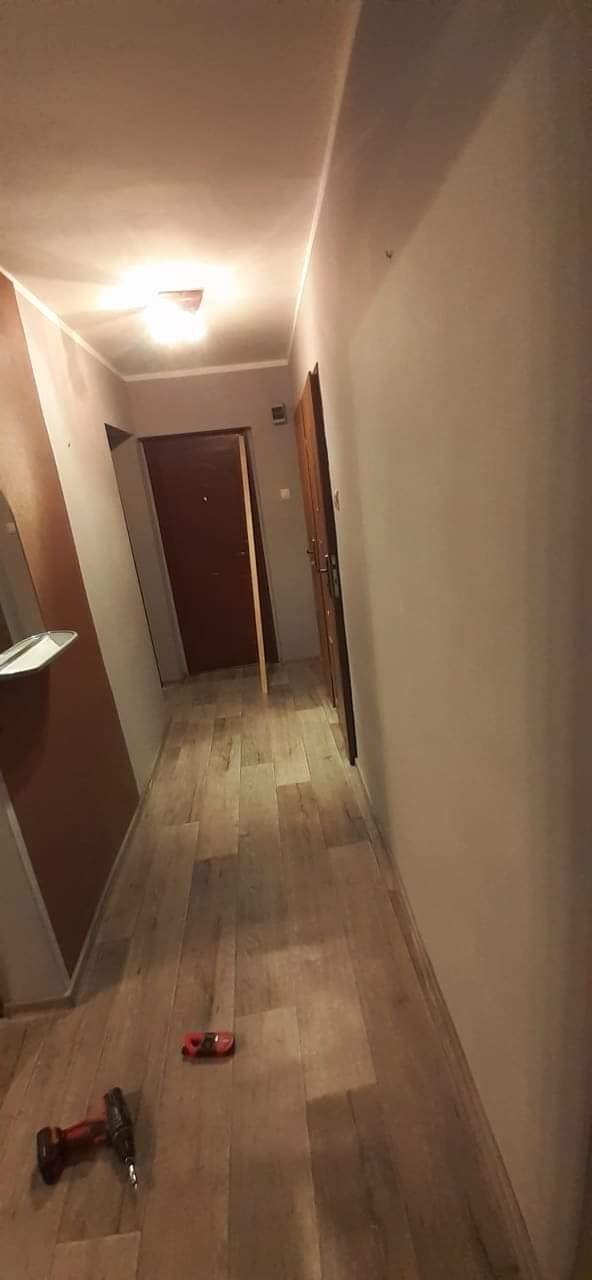 Korytarz po remoncie: świeżo pomalowane ściany w odcieniach szarości i położona podłoga z paneli imitujących drewno; w tle widoczne drzwi i narzędzia na podłodze.