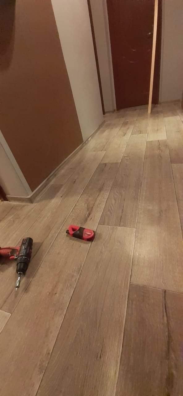 Montaż paneli podłogowych z użyciem wkrętarki i docinaka, widoczna brązowa ściana w tle.