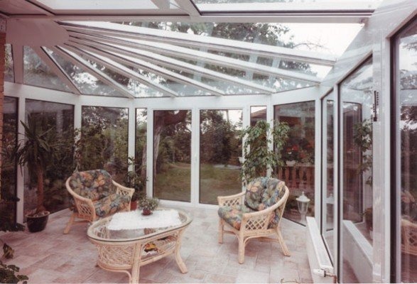 Jasna oranżeria z białymi ramami okiennymi i szklanym dachem, wyposażona w wiklinowe meble wypoczynkowe i rośliny doniczkowe, z widokiem na ogród.