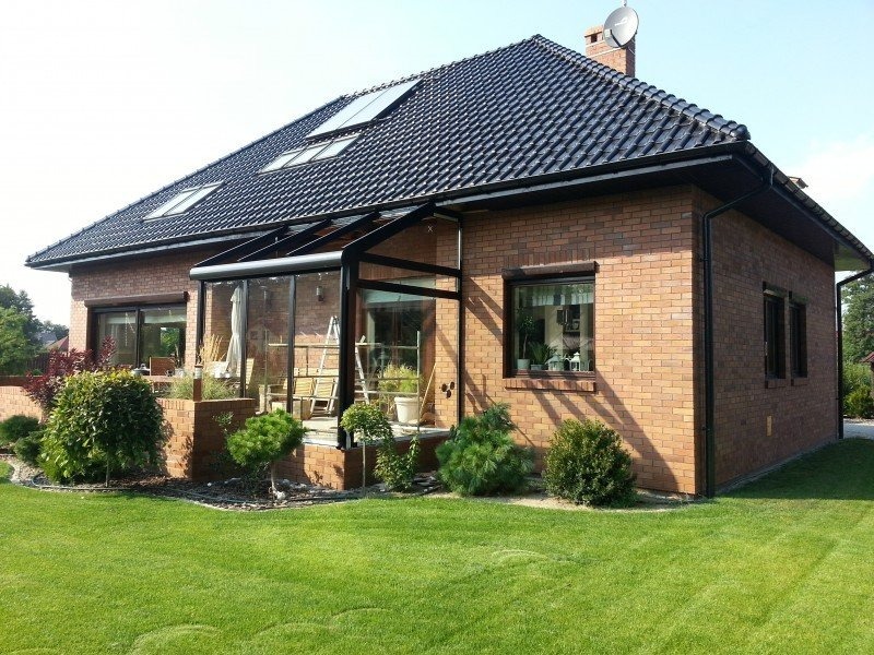 Dom z cegły z przeszkloną werandą z czarną ramą, widok z zewnątrz na zadbany trawnik i dekoracyjne rośliny.