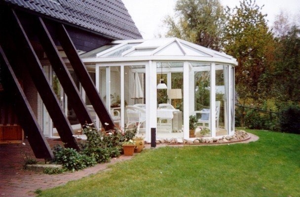 Ogród zimowy z białymi ramami szklanymi, widoczny z zewnątrz, z elementami konstrukcyjnymi domu w tle i zielonym trawnikiem.