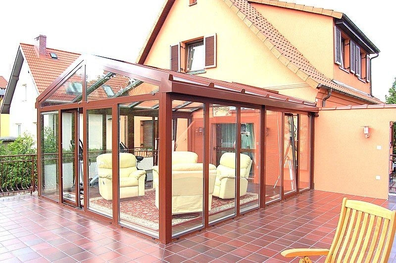 Zadaszony taras z brązowymi ramami, przeszklonymi ścianami i dachem, z widocznymi beżowymi fotelami wypoczynkowymi wewnątrz oraz czerwono-brązową terakotą na podłodze tarasu.