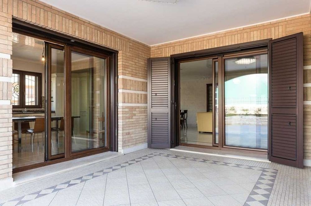 Zewnętrzny taras z przesuwnymi, brązowymi drzwiami balkonowymi z PCV i drewnianymi okiennicami, wykończony cegłą i płytkami podłogowymi w geometryczny wzór.