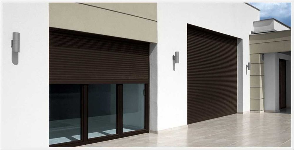 Elewacja domu z brązowymi roletami zewnętrznymi opuszczonymi na oknach i bramie garażowej, oświetlona kinkietami w kształcie walca.