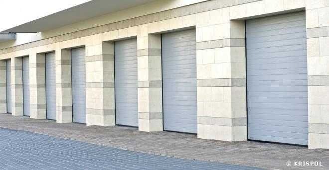 Szereg zamkniętych, szarych bram garażowych segmentowych w jasnej, kamiennej elewacji budynku.