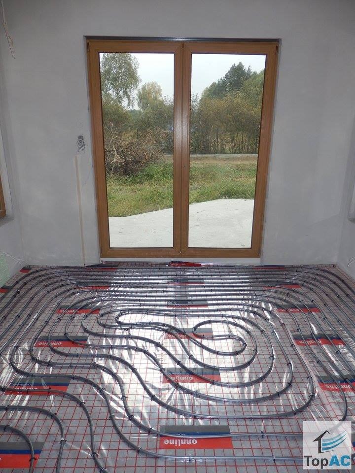 Instalacja ogrzewania podłogowego w surowym wnętrzu z widokiem na zewnątrz przez duże okno balkonowe.