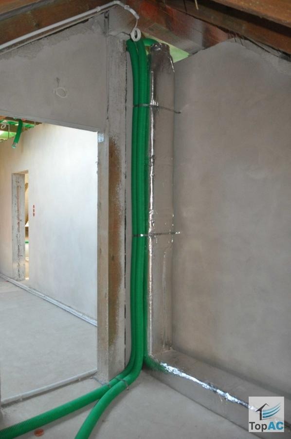 Surowe wnętrze w trakcie prac wykończeniowych; widoczne pionowe rury wentylacyjne w zielonej karbowanej osłonie, przymocowane do ściany za pomocą metalowych opasek, fragment izolacji termicznej...