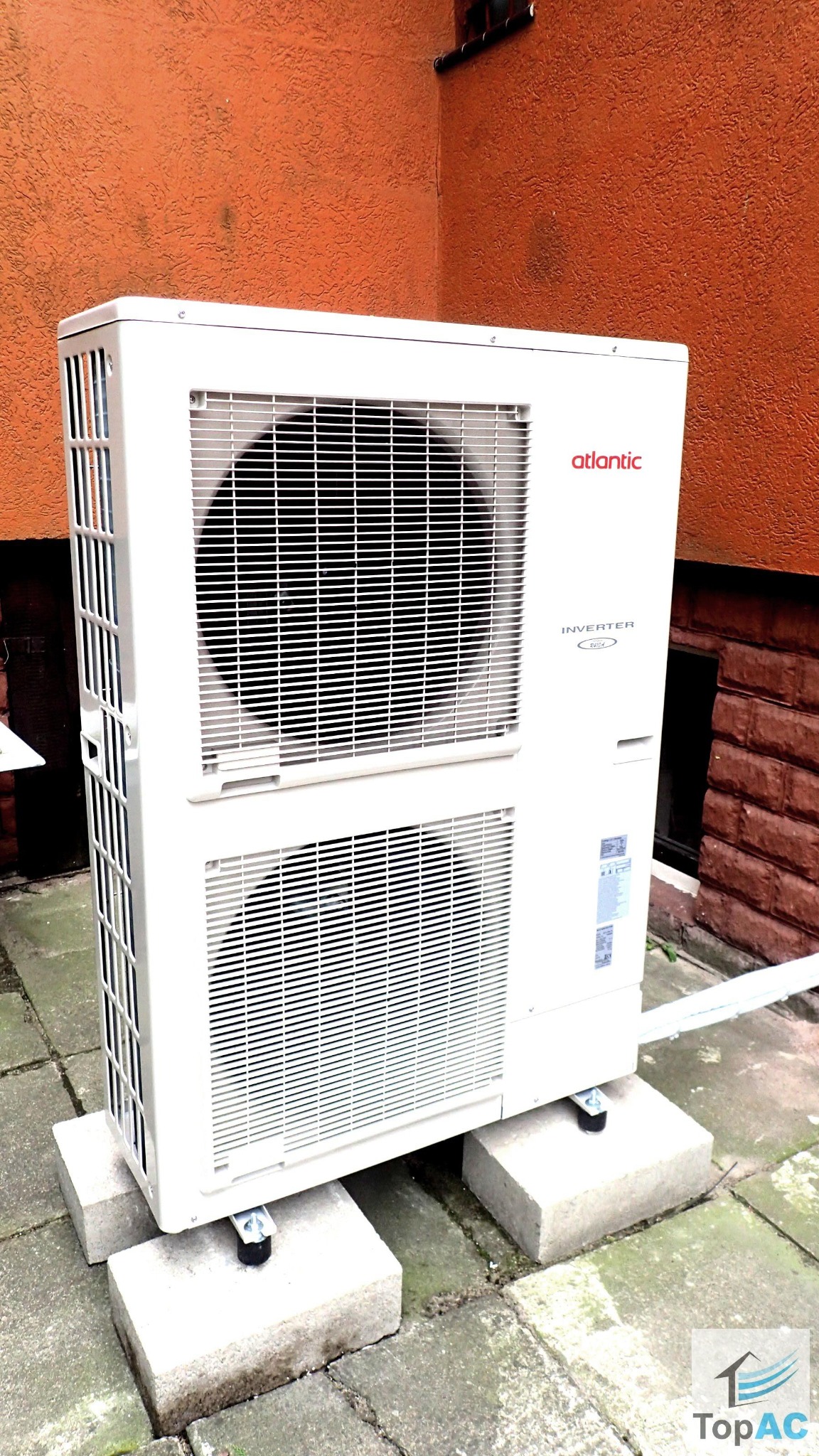 Biała jednostka zewnętrzna klimatyzacji marki Atlantic, umieszczona na betonowych blokach na tle pomarańczowej ściany z ceglanym fragmentem.