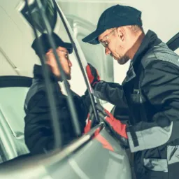 Mężczyzna w okularach i roboczym kombinezonie, w czerwonych rękawicach, montuje lub naprawia szybę w samochodzie. Widoczne odbicie mechanika w szybie.