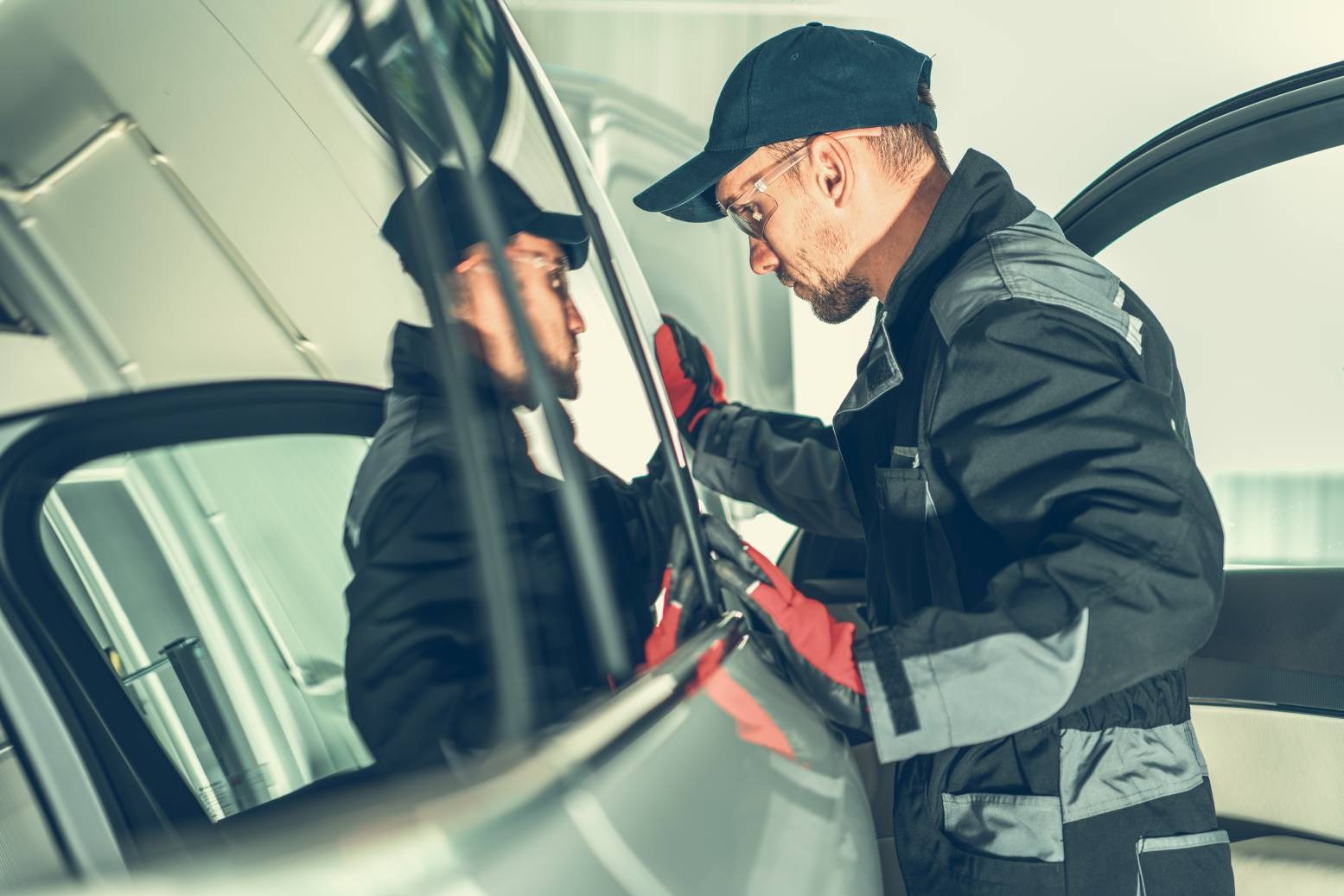 Mężczyzna w okularach i roboczym kombinezonie, w czerwonych rękawicach, montuje lub naprawia szybę w samochodzie. Widoczne odbicie mechanika w szybie.