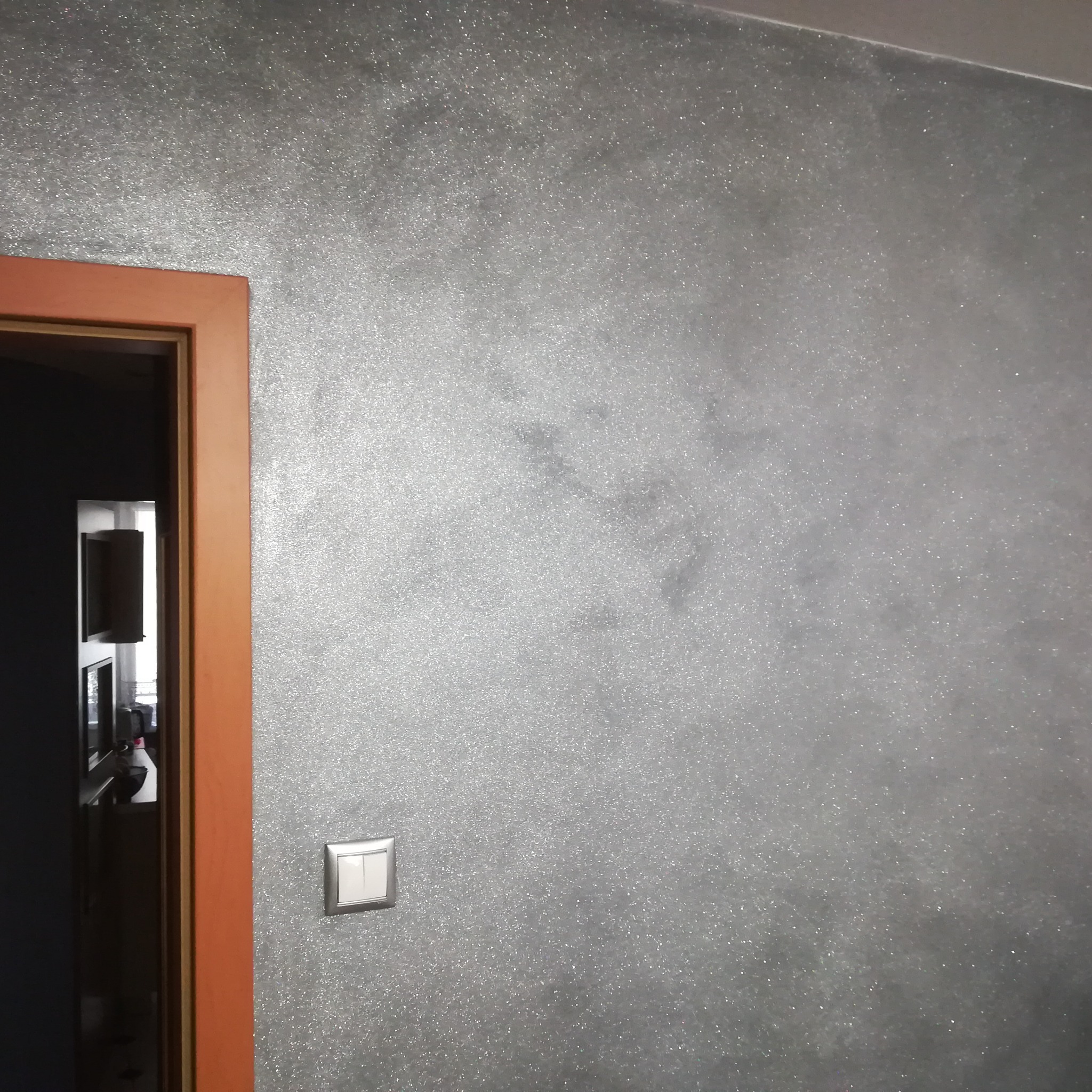 Ściana w odcieniach szarości z efektem brokatu, widoczny włącznik światła i fragment futryny drzwi w kolorze drewna.