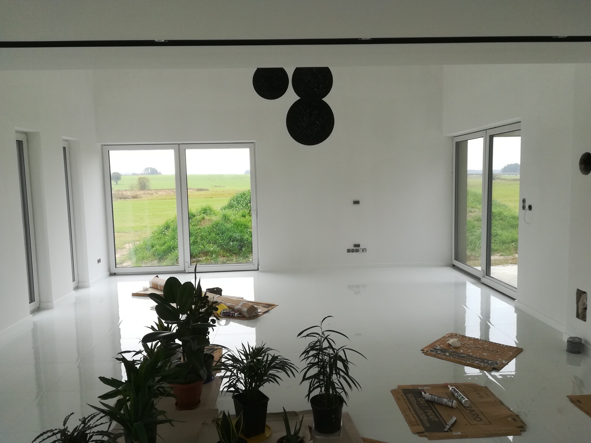 Białe, minimalistyczne wnętrze z dużymi oknami wychodzącymi na zielony krajobraz. Podłoga pokryta błyszczącą wylewką. Na podłodze kartony zabezpieczające i rośliny doniczkowe. Na ścianie dekoracja...