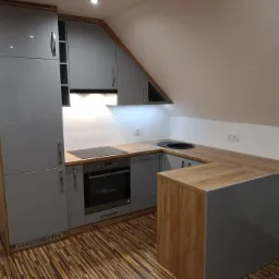 Szara kuchnia z drewnianymi akcentami, wkomponowana w przestrzeń pod skosem dachu, z widoczną płytą indukcyjną i okrągłym zlewem.