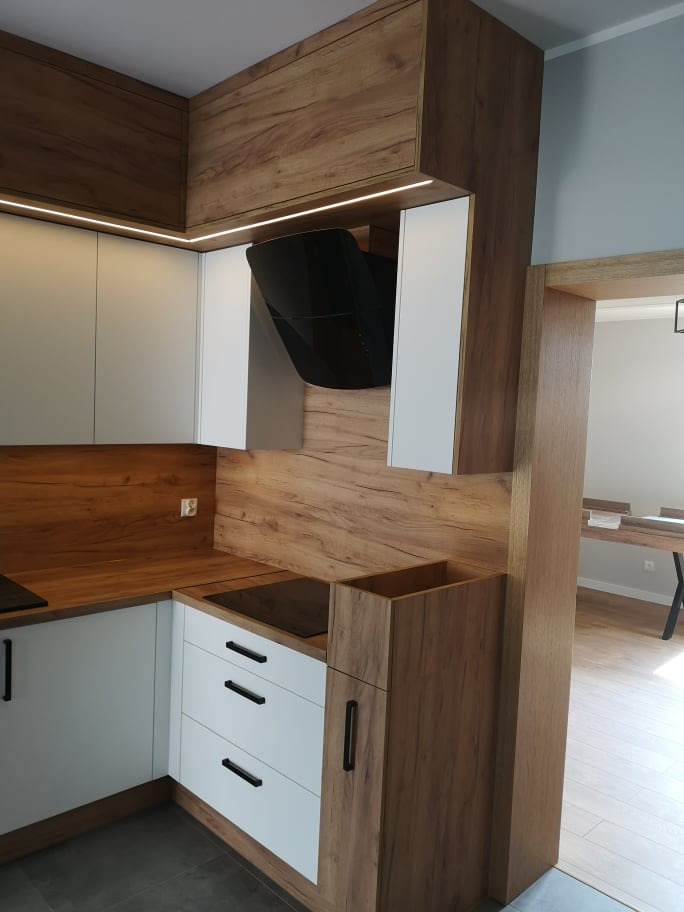 Nowoczesna kuchnia z białymi szafkami dolnymi i górnymi, drewnopodobnymi blatami i obudową, czarnym okapem i listwami LED pod szafkami wiszącymi.