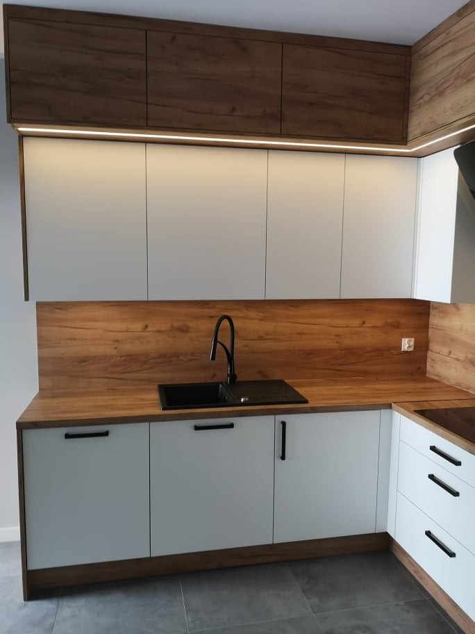 Nowoczesna kuchnia z białymi szafkami dolnymi i górnymi z drewnianymi akcentami, czarnym zlewozmywakiem i baterią, podświetleniem LED pod górnymi szafkami i drewnianym panelem nad blatem.