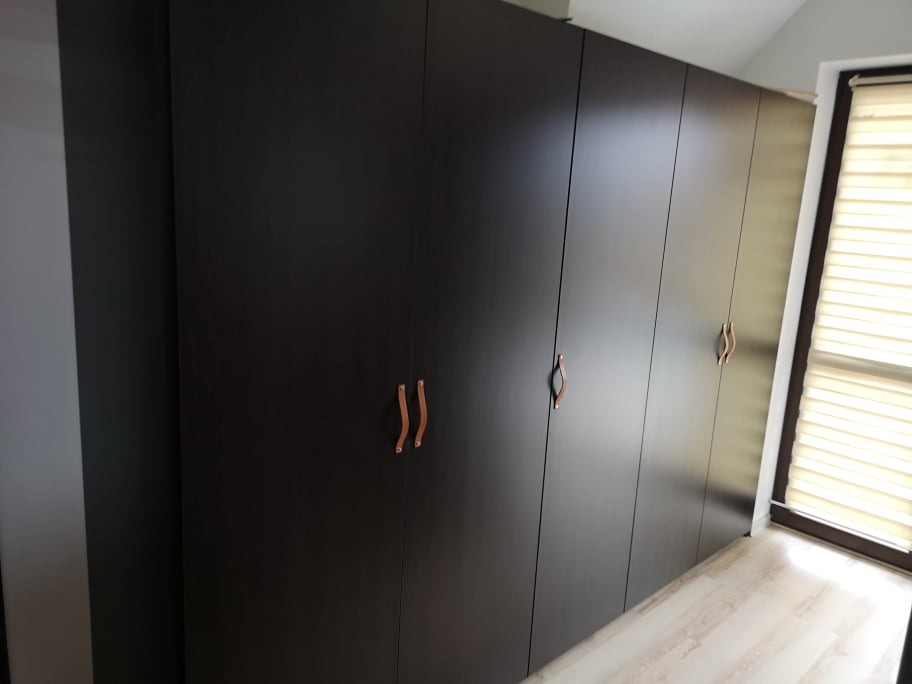 Ciemna, wysoka zabudowa szafowa z minimalistycznymi, skórzanymi uchwytami w kolorze brązowym, widoczna w jasnym wnętrzu przy oknie z żaluzjami.