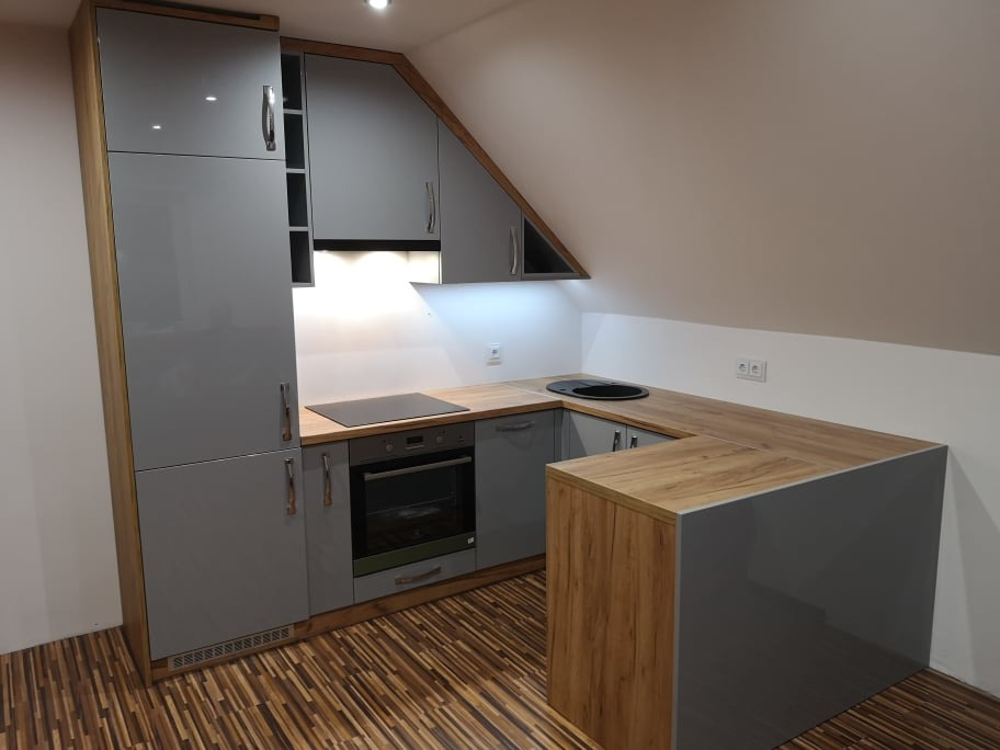 Szara kuchnia z drewnianymi akcentami, wkomponowana w przestrzeń pod skosem dachu, z widoczną płytą indukcyjną i okrągłym zlewem.